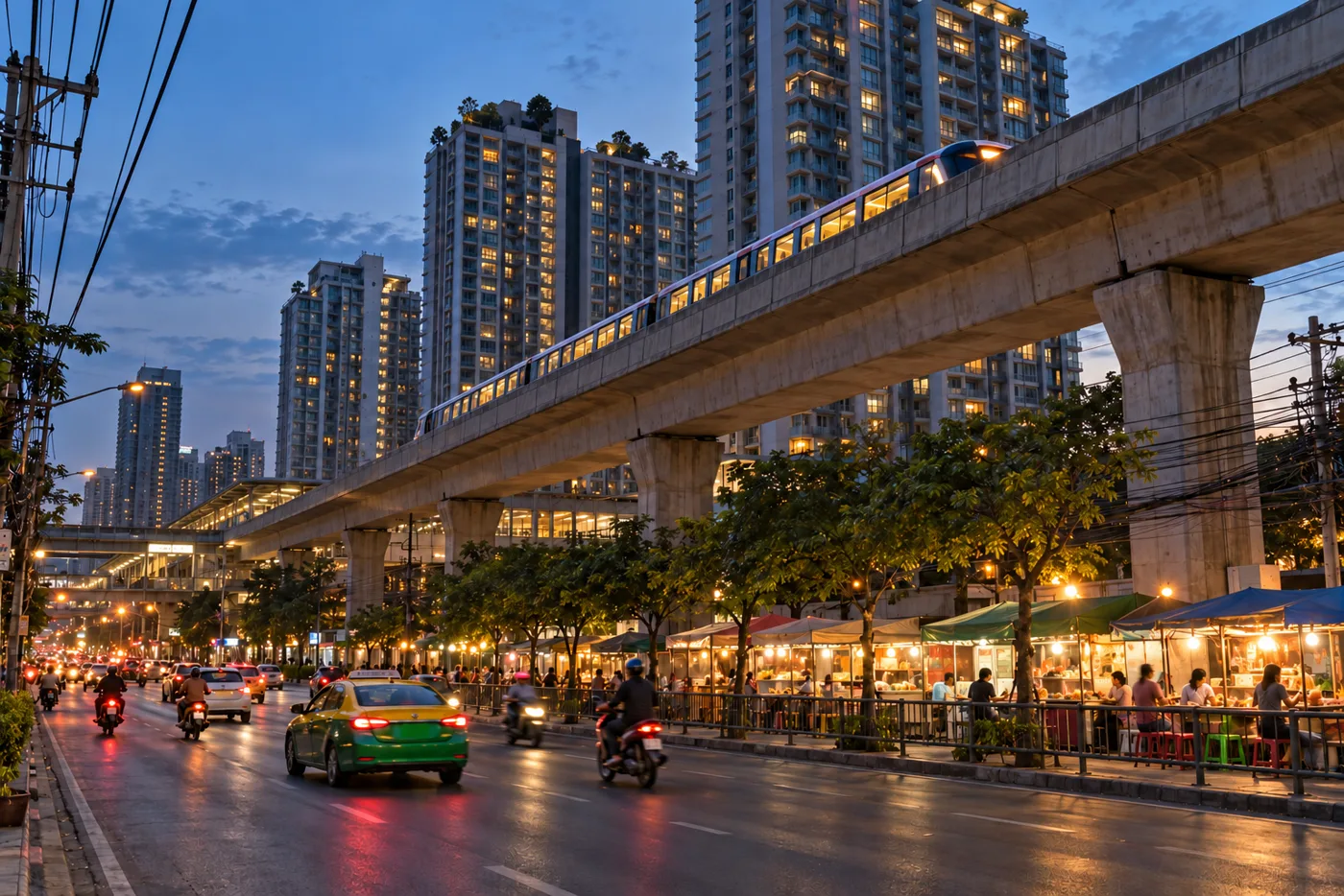 Early evening at On Nut in Bangkok, with the elevated Skytrain track running above a busy road, condominium towers behind it and food stalls lit along the pavement