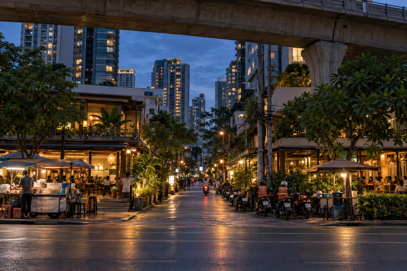A busy side street off Sukhumvit Road in Bangkok at dusk, with the BTS viaduct overhead, street food carts on the pavement and condominium towers lit up behind.