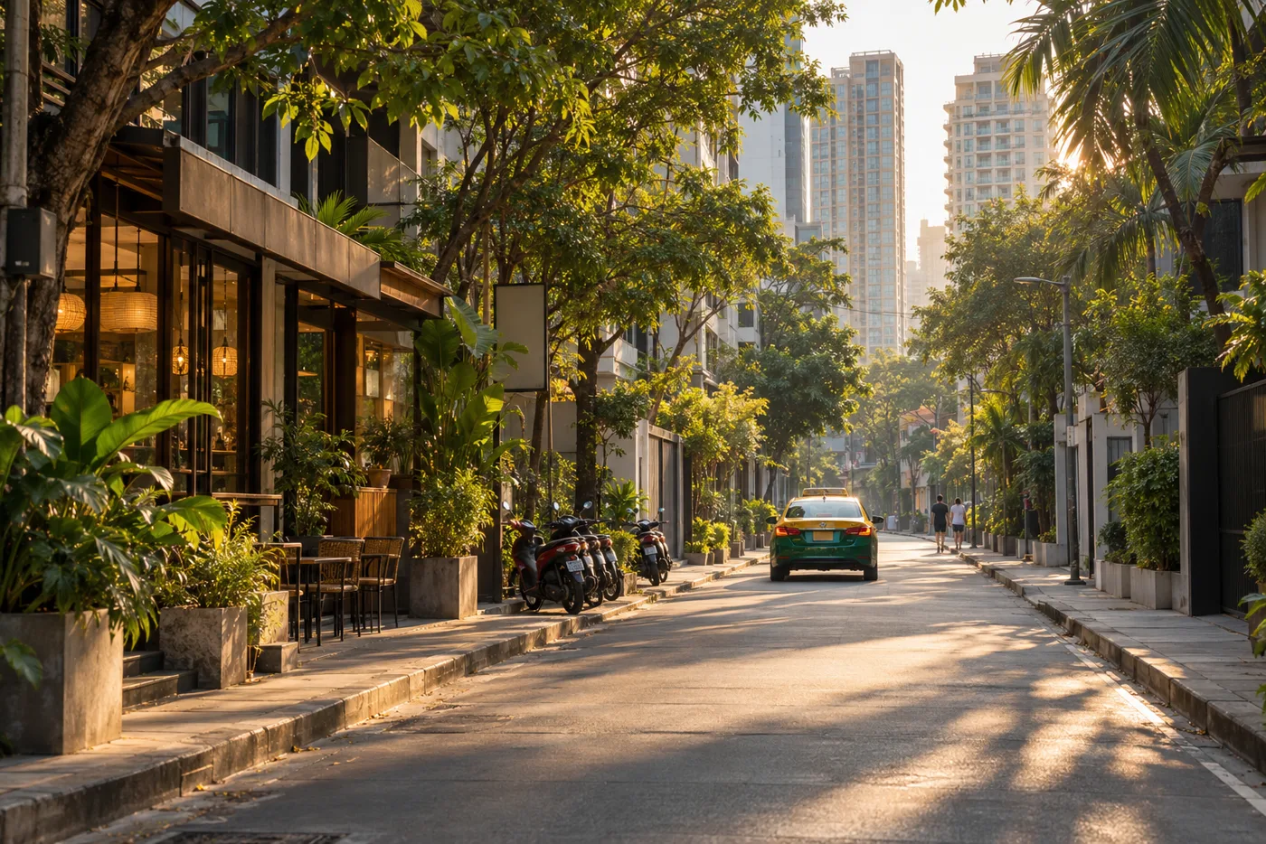 A long, tree-lined side street off Sukhumvit Road in Bangkok in the late afternoon, with low shophouses, cafés and tall residential towers on both sides and a few motorbikes parked along the kerb.