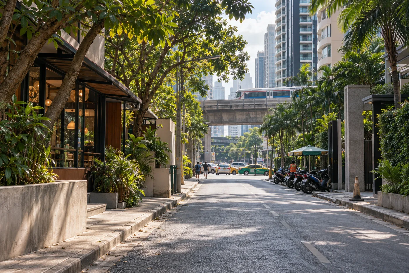A tree-lined side street off Sukhumvit Road in Bangkok in the morning, with condominium towers on both sides and the BTS Skytrain viaduct crossing the far end of the street.