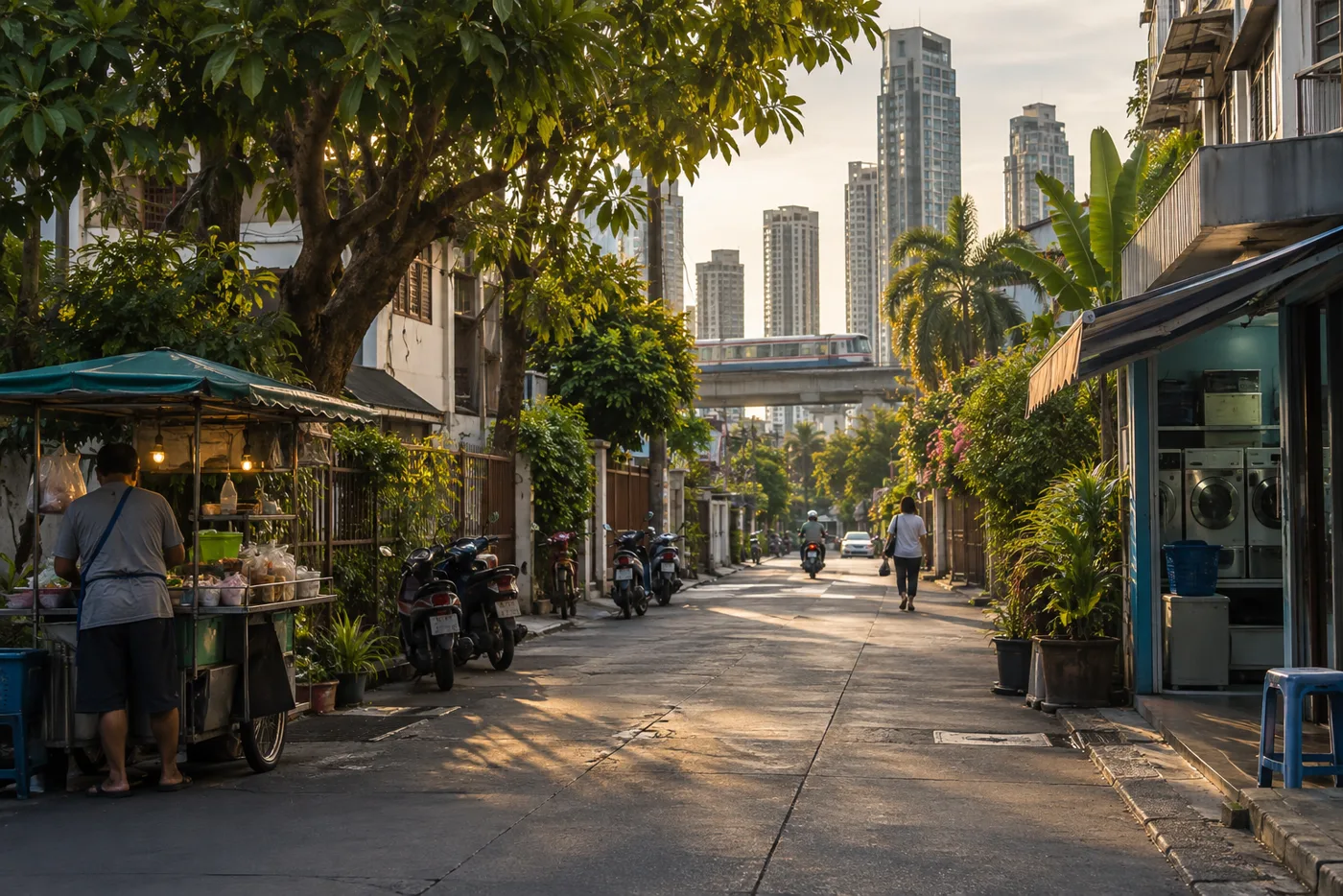 A residential street of condominium towers in central Bangkok in the late afternoon, with street trees, food carts on the pavement and an elevated railway line in the distance