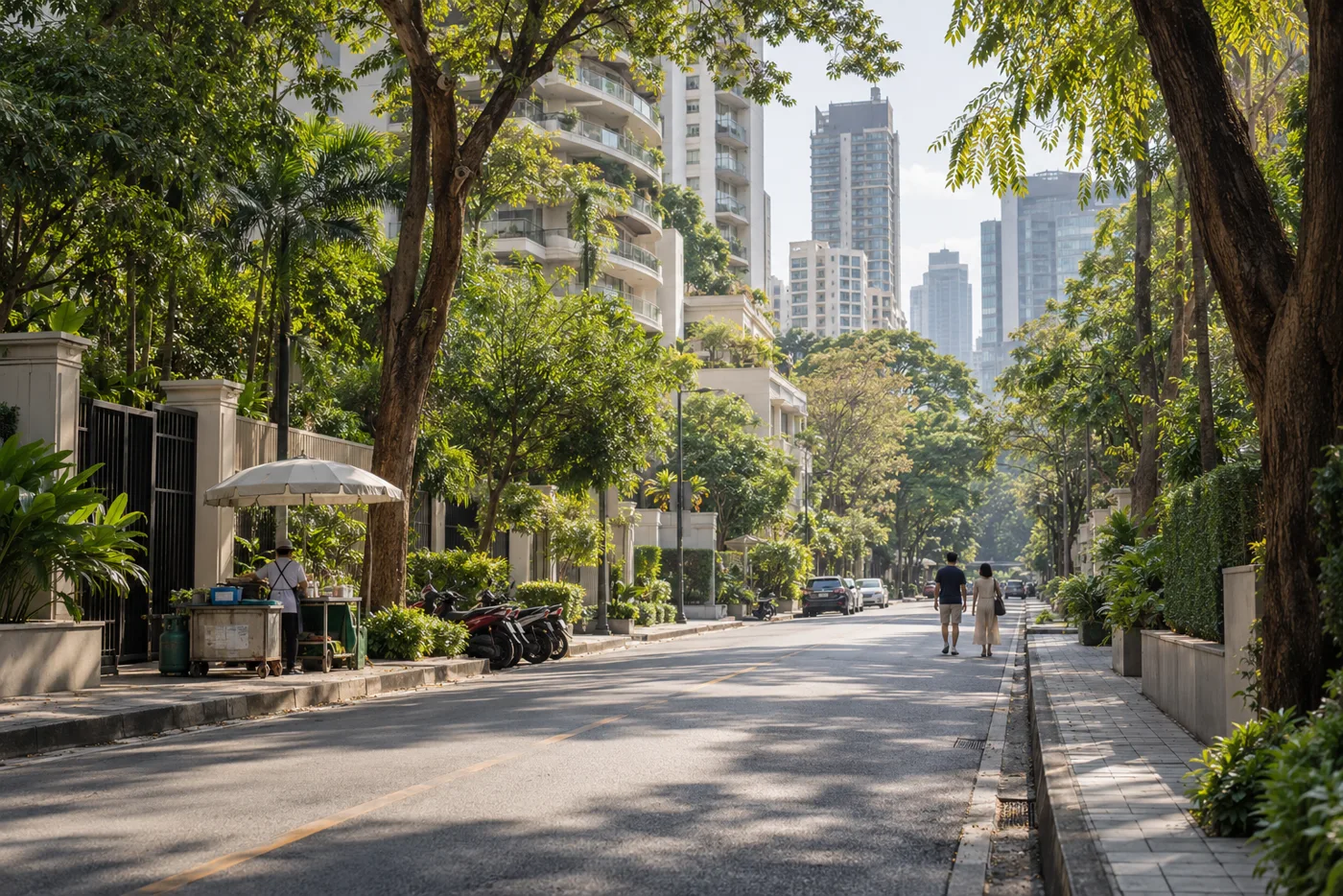 A tree-lined side street in central Bangkok in the morning, with tall residential towers rising behind the trees and a few motorbikes parked along the pavement.