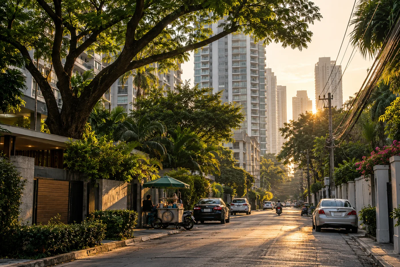 A tree-lined residential lane in central Bangkok in late-afternoon light, with tall condominium towers and their balconies behind the trees and a street-food cart on the pavement