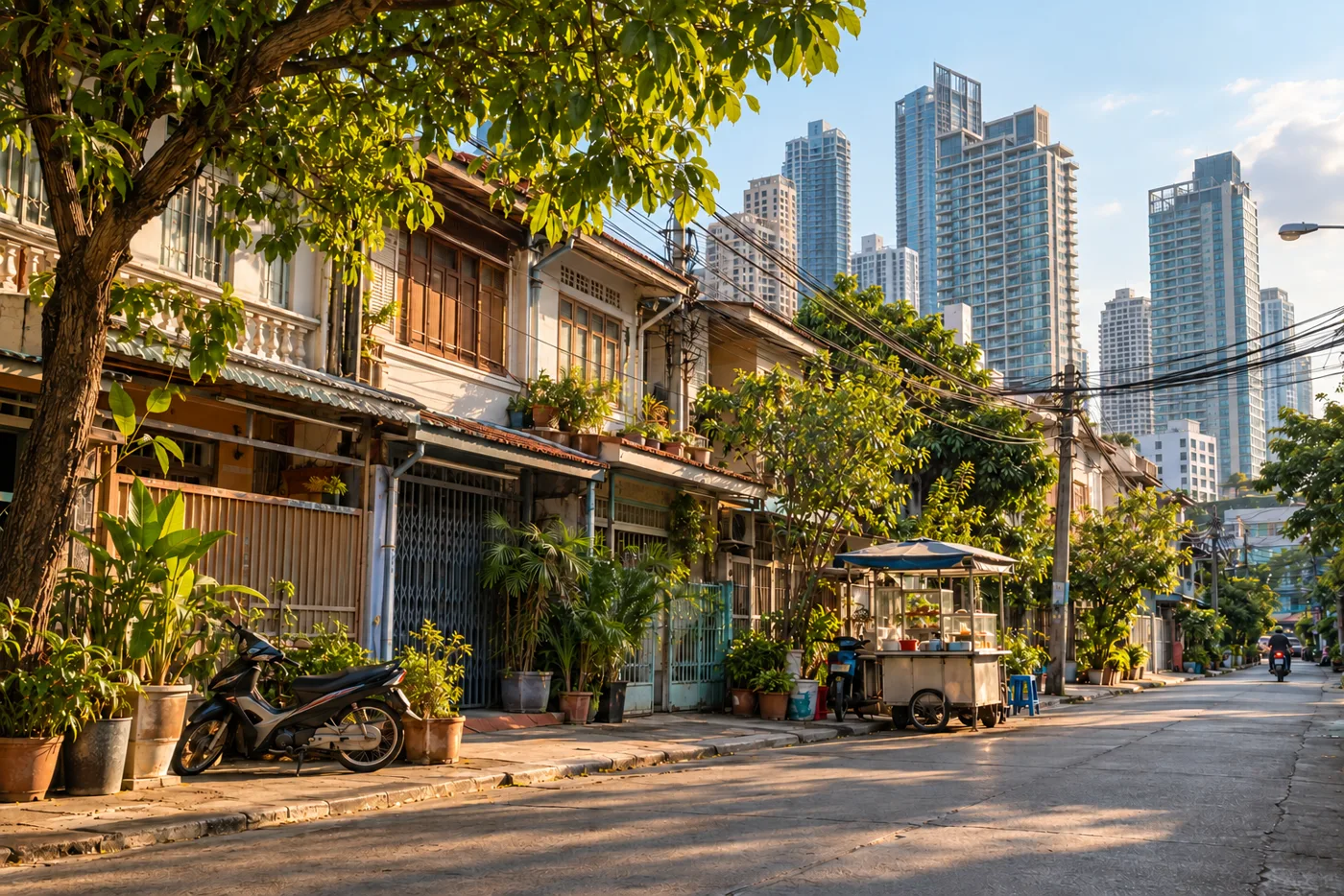 A quiet residential soi in Bangkok in the late afternoon, with two-storey townhouses and potted plants in the foreground and tall condominium towers rising behind them
