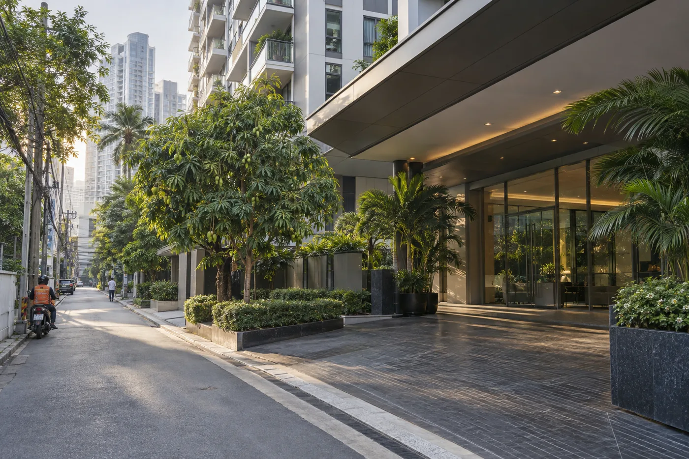 The ground-floor entrance of a Bangkok condominium tower on a leafy residential soi, with a covered driveway and glass doors leading into a lobby