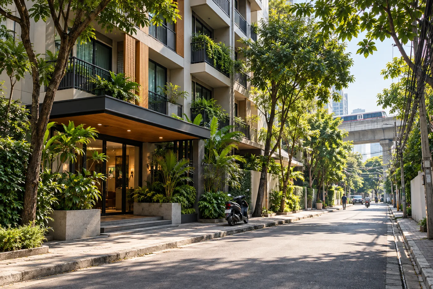 A quiet residential soi in Bangkok in the morning, with a mid-rise apartment building, potted plants by the entrance and the elevated Skytrain line at the end of the street