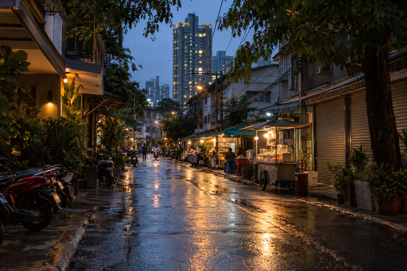 A narrow Bangkok side street off Sukhumvit Road in the early evening, food stalls and low shophouses on one side and a tall condominium tower rising behind them